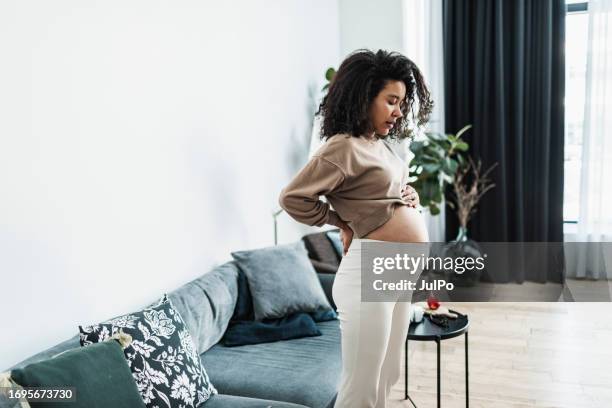 schwangere schwarze frau sitzt auf dem sofa und leidet unter rückenschmerzen, müde von der arbeit - symptom stock-fotos und bilder
