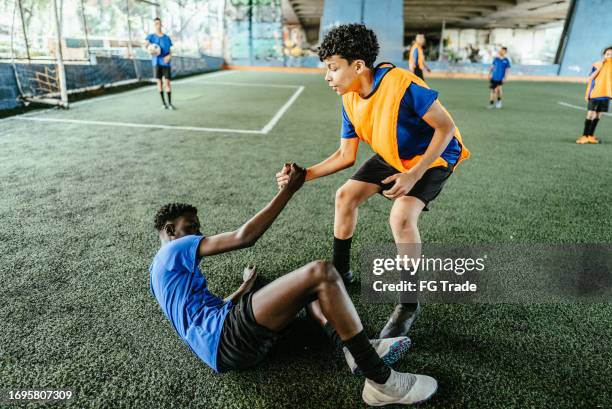fußballspieler hilft dem gegnerischen spieler, auf das fußballfeld zu kommen - fairness stock-fotos und bilder