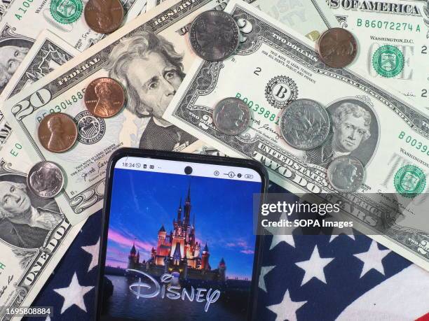 In this photo illustration, The Walt Disney Company logo seen displayed on a smartphone with United States Dollar notes and coins in the background.