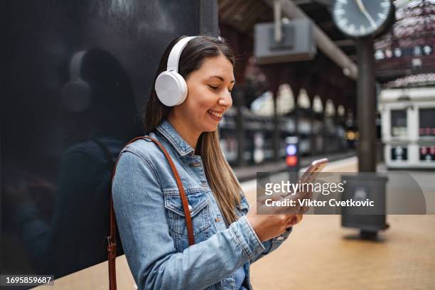 young woman with wireless headphones listening to music and texting on the phone while waiting for her train at the train station - copenhagen metro stock pictures, royalty-free photos & images