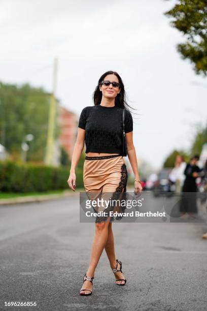 Gala Gonzalez wears a black t-shirt with printed monograms, a beige silk lustrous on-knee skirt with black lace / embroidery, Gucci shoes, outside...