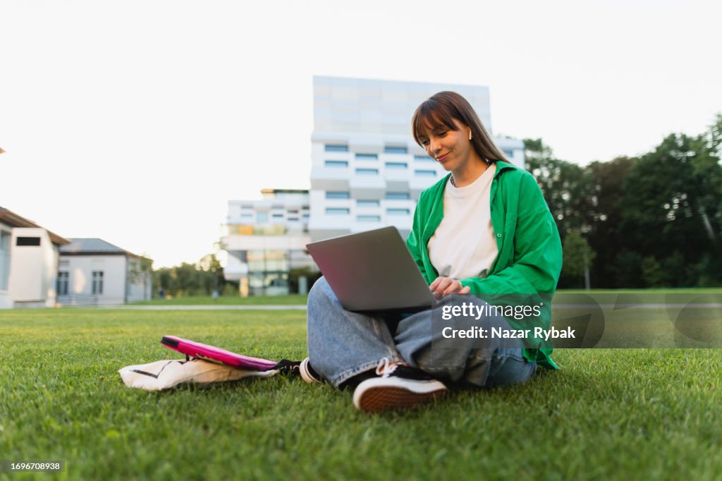A beautiful young woman studying at the laptop sitting on the grass on the background of the campus