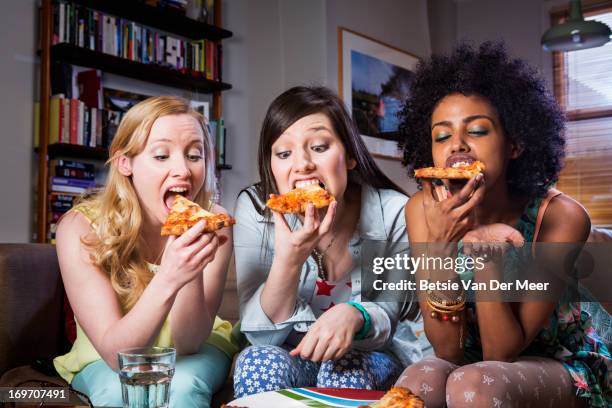 women biting into pizza sitting together on sofa. - comer pizza imagens e fotografias de stock