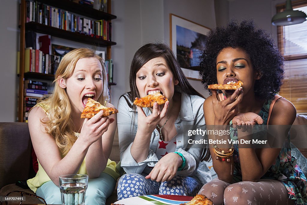 Women biting into pizza sitting together on sofa.