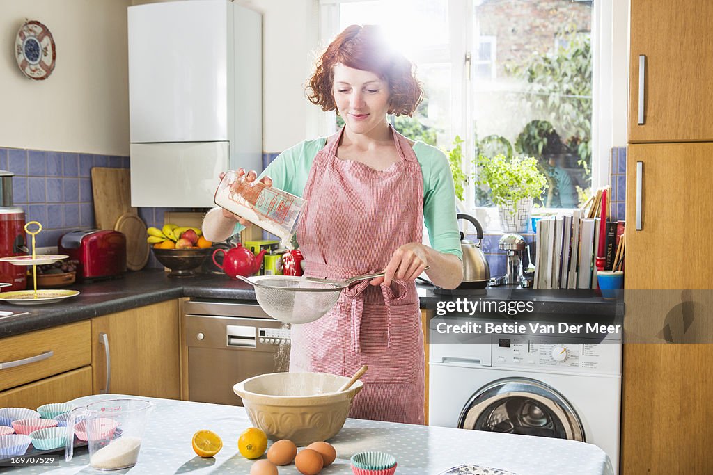 Woman sieving flour in bowl in kitchen.