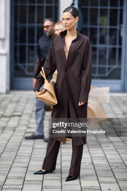 Bianca Balti is seen wearing gold hoop earrings, black shiny pumps, a light beige leather Ferragamo bag with gold hardware and a brown long shirt...