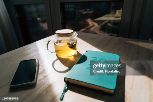 a blue diary for 2024 on a sunlit table, a teapot with yellow herbal tea and a mobile phone - diet journal stockfoto's en -beelden