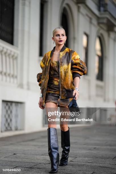 Caroline Daur wears earrings, a yellow and brown leather tie and dye bomber jacket, a gathered cropped top, matching mini skirt, a watch, a black...