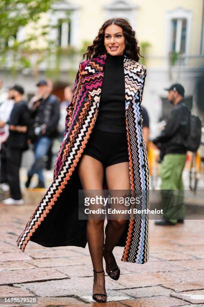Paola Turani wears a black turtleneck ribbed top, mini shorts, a purple / orange and blue geometric pattern print long trench coat, tights, high...