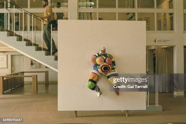 Gallery visitor walks up a flight of stairs behind a piece of work, a multi-coloured figure mounted on a white board on display as part of 'Hon - en...