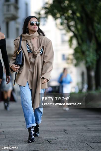 Guest wears sunglasses, a beige scarf, beige oversized blazer jacket, black and white checked bras, blue denim jeans / pants with hems, black shiny...
