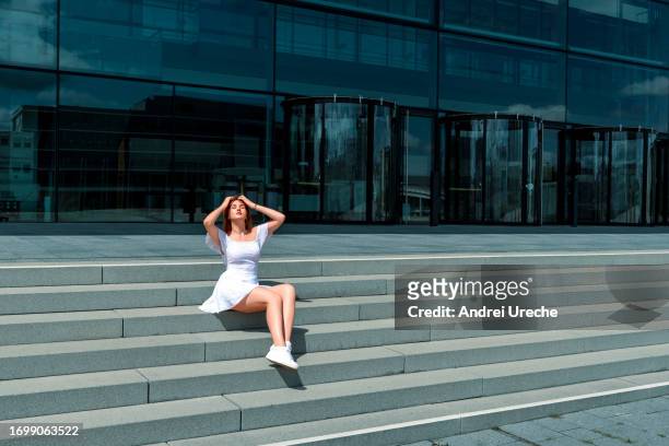 woman in elegant white dress sitting on stairs in front of glass building - sneakers street style stock pictures, royalty-free photos & images