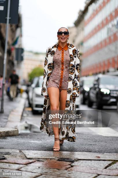 Ginevra Mavilla wears sunglasses, earrings, a white and brown floral print long coat, an orange and brown shirt worn as mini / short ribbed dress...