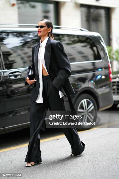 Izabel Goulart wears sunglasses, a white shirt, a black oversize blazer jacket, flowing matching pants, sandals, outside Dolce & Gabbana, during the...