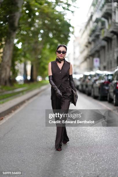 Guest wears suglasses, a choker with flower detail, a brown full look, waistcoat, leather long gloves, a clutch, suit flared pants, outside Dolce &...