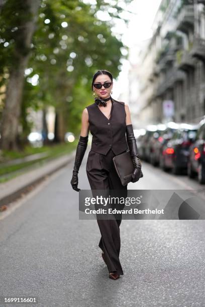 Guest wears suglasses, a choker with flower detail, a brown full look, waistcoat, leather long gloves, a clutch, suit flared pants, outside Dolce &...