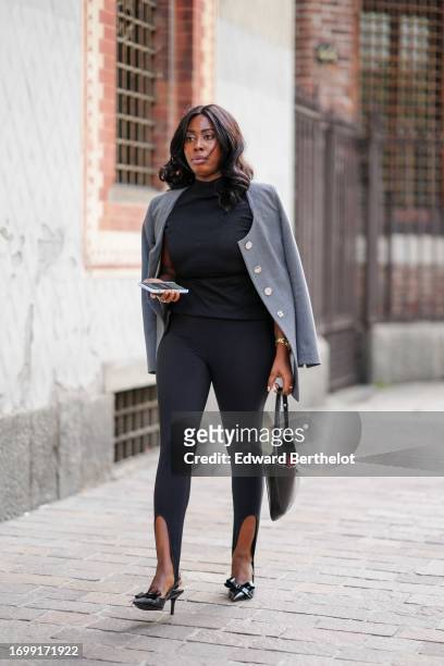 Guest wears a black turtleneck top, black slim stirrup leggings, a black bag, black pointed stilettos with a bow, a grey jacket with white buttons,...