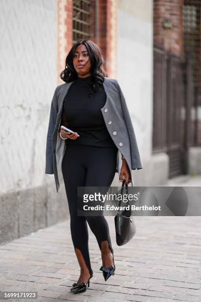 Guest wears a black turtleneck top, black slim stirrup leggings, a black bag, black pointed stilettos with a bow, a grey jacket with white buttons,...