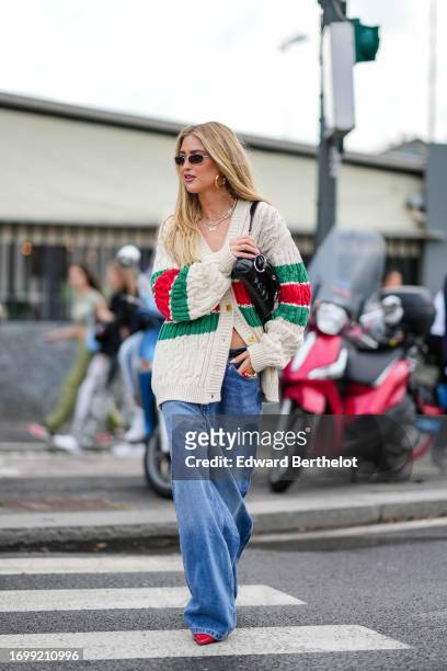 Emili Sindlev wears sunglasses, earrings, a necklace, a green red and white striped / logo wool knit cardigan, a black leather bag, blue denim...
