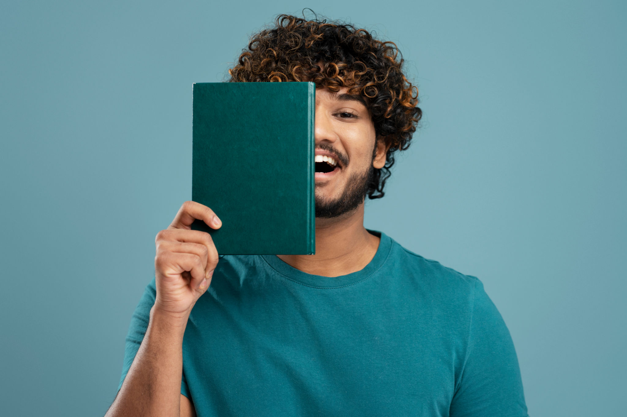 Cheerful Indian male student smiling and covering half of his face with a green hardcover book Cheerful Indian male student smiling and covering half of his face with a green hardcover book
