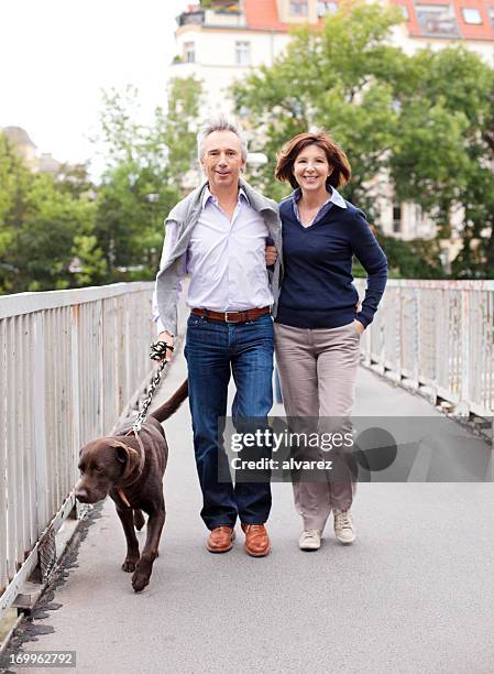 mature adult couple walking the dog - mid adult couple stockfoto's en -beelden
