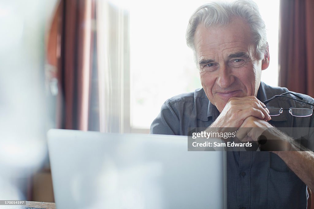 Portrait of smiling senior man holding eyeglasses and using laptop