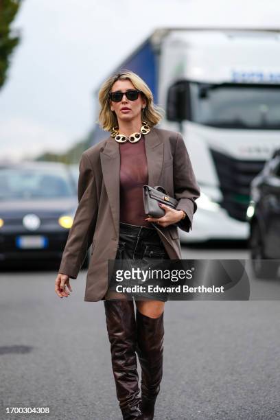 Guest wears sunglasses, a large golden necklace, a plum color top, a light brown jacket, a black leather mini skirt with large pockets, brown leather...
