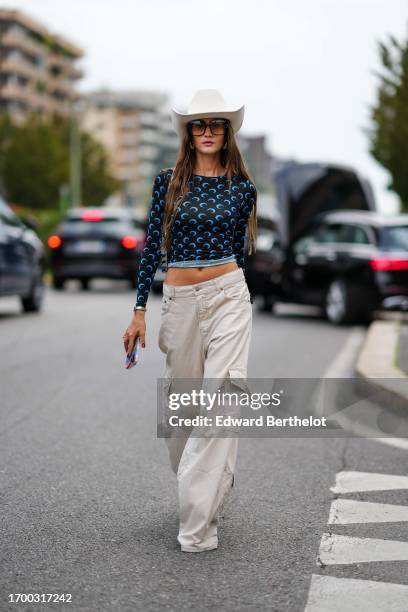 Guest wears a white cowboy hat, a black long-sleeved top with a blue pattern, white cargo jeans, outside Anakiki, during the Milan Fashion Week -...