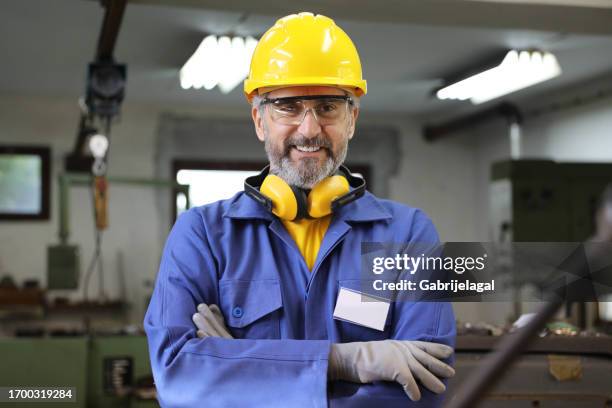 a close-up view of a handsome mid-50s factory worker posing for the camera at his workplace. he is standing in front of the machine he is driving, looking at the camera with folded arms and smiling. - employee of the month stock pictures, royalty-free photos & images