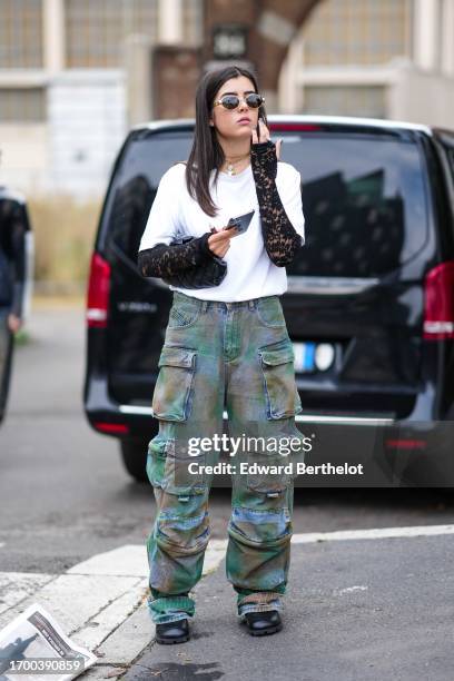 Guest wears sunglasses, necklaces, a white top, black lace mittens, camouflage cargo pants, black leather shoes, outside Anakiki, during the Milan...