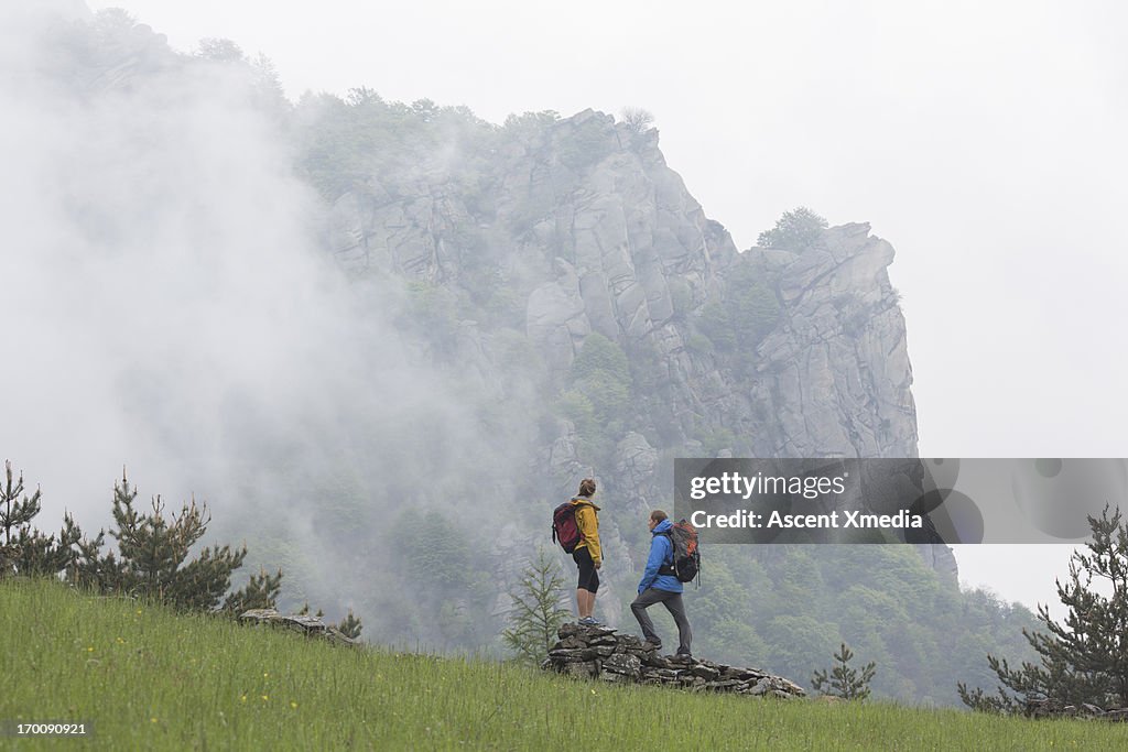 Hiker couple stand on rock wall, watch mist swirl