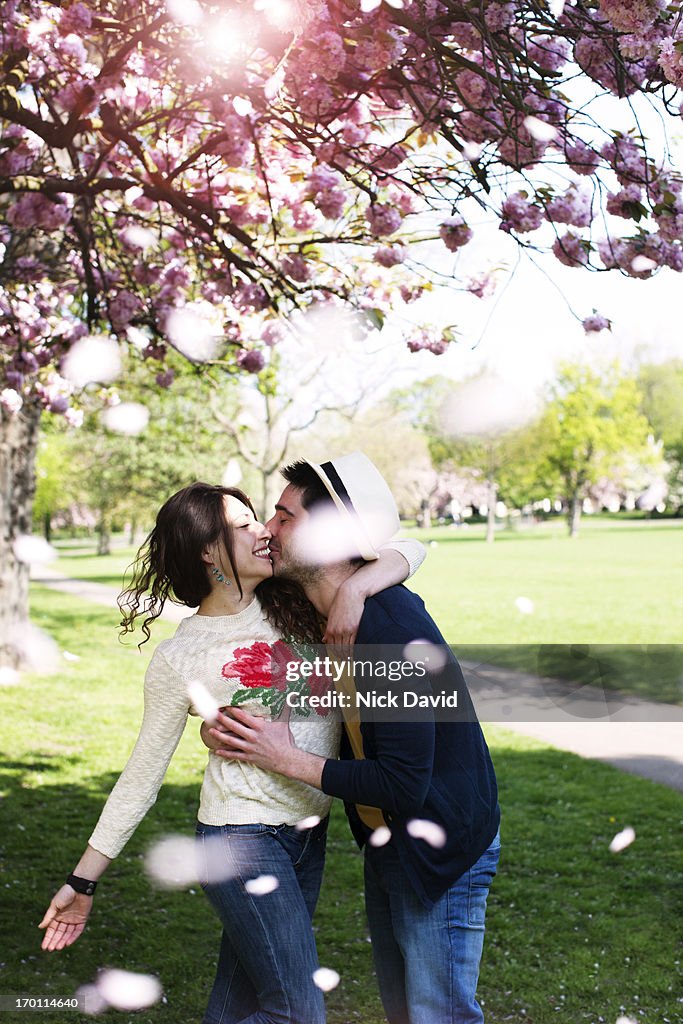 A young couple embrace and kiss in a sunny park under a pink blossom tree