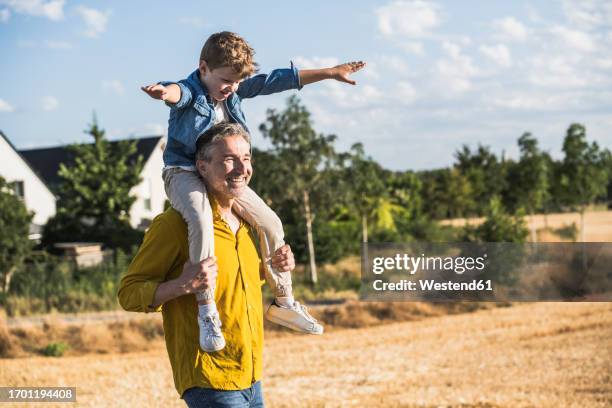 cheerful grandfather carrying grandson on shoulders in field - person auf den schultern tragen stock-fotos und bilder