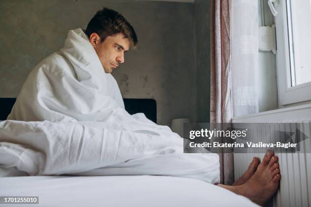 man tries to warm his feet on a radiator while sitting under a blanket on the bed - emmitouflé dans une couverture photos et images de collection
