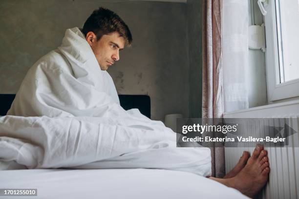 man tries to warm his feet on a radiator while sitting under a blanket on the bed - frozen man stockfoto's en -beelden