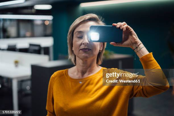businesswoman covering eye with mobile phone and flashing light in office - iphone flashlight stock pictures, royalty-free photos & images