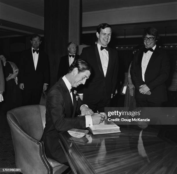 British Royal Charles, Prince of Wales, wearing a tuxedo and bow tie, smiles as he sits signing a guest book, with two men laughing beside the table...