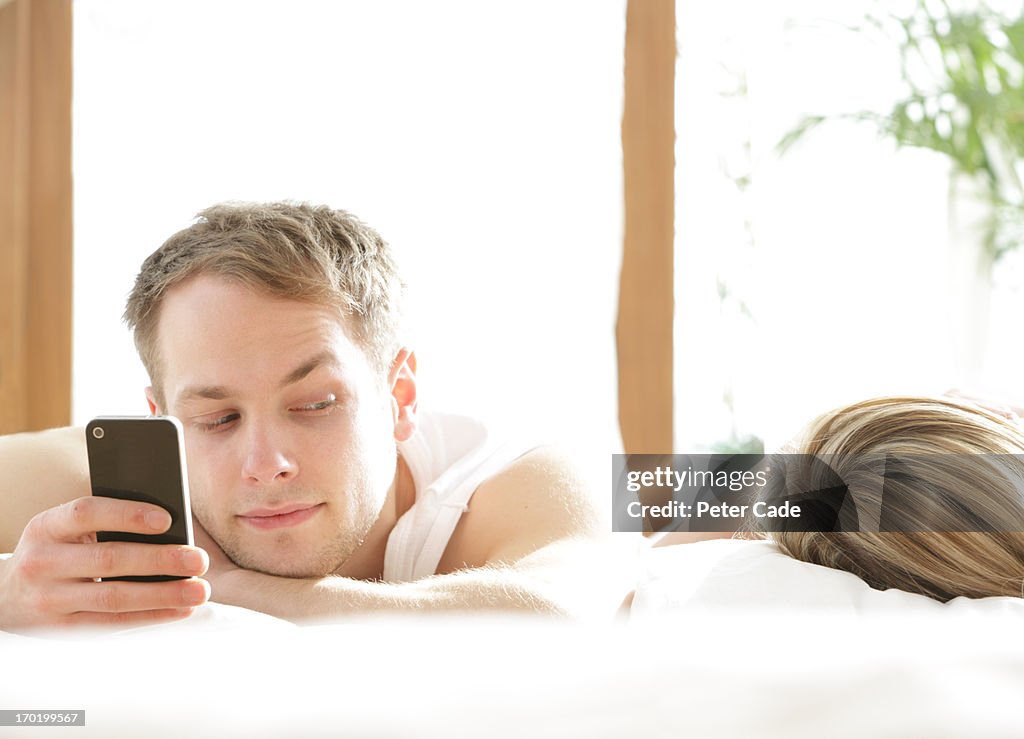 Man with phone in bed, looking at woman asleep