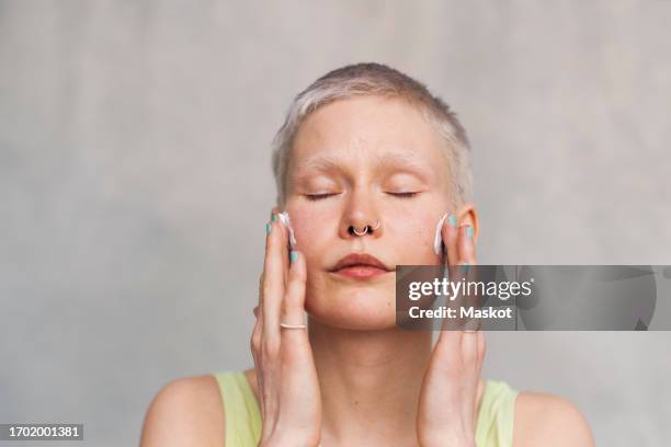 woman with eyes closed applying face cream against white background - gezichtscrème stockfoto's en -beelden