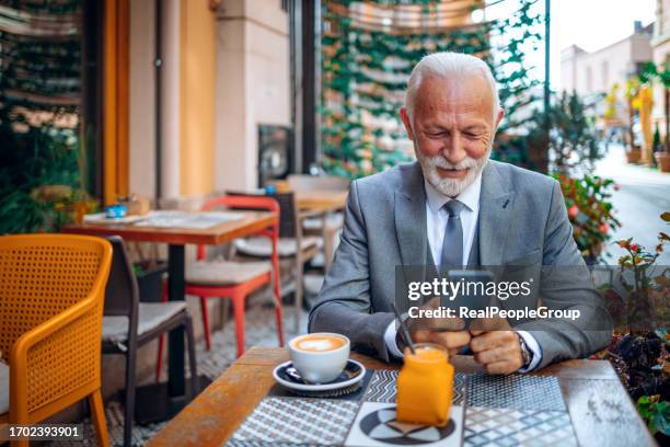 ein älterer geschäftsmann, der fröhlich auf sein mobiltelefon schaut, während er in einem örtlichen café sitzt - real businessman isolated no smile stock-fotos und bilder