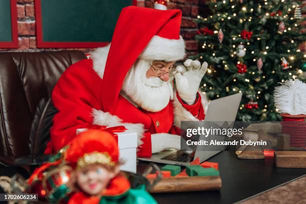 santa claus using a laptop while sitting at a table against the background of toys and a christmas tree. - santa hat and beard stock pictures, royalty-free photos & images