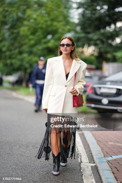Guest wears sunglasses, earrings, an oversized jacket, a black macrame fringe dress, a red bag, black leather cowboy boots decorated with rhinestone...