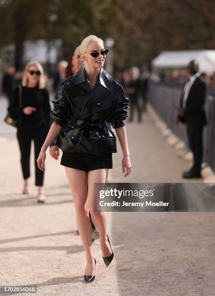 Anya Taylor-Joy is seen wearing a complete outfit from Christian Dior, consisting of black cat eye sunglasses; a waisted, shiny, black leather jacket...