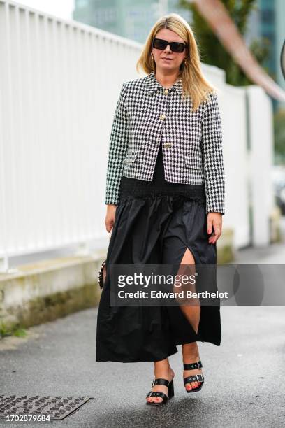 Guest wears sunglasses, earrings, a black and white houndstooth jacket, a black ruffled side-slit long skirt, black sandals, outside Moschino, during...