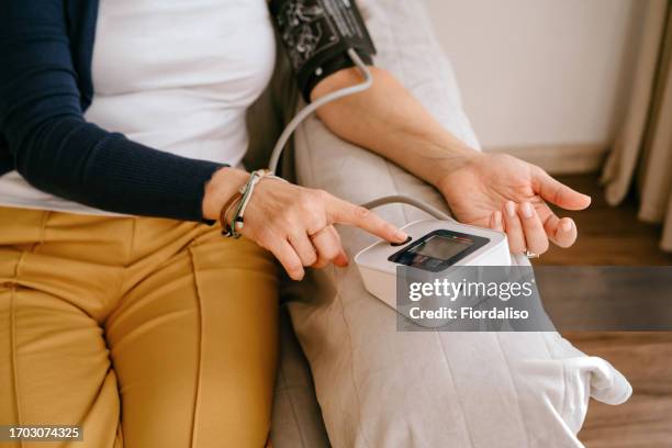 a middle-aged woman sitting on the sofa, using a tonometer to measure blood pressure - medidor de tensão arterial imagens e fotografias de stock