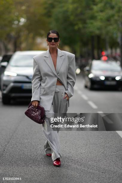 Gili Biegun wears sunglasses, earrings, a gray oversized blazer jacket, a burgundy leather bag, pointed shoes, outside Anrealage, during the...
