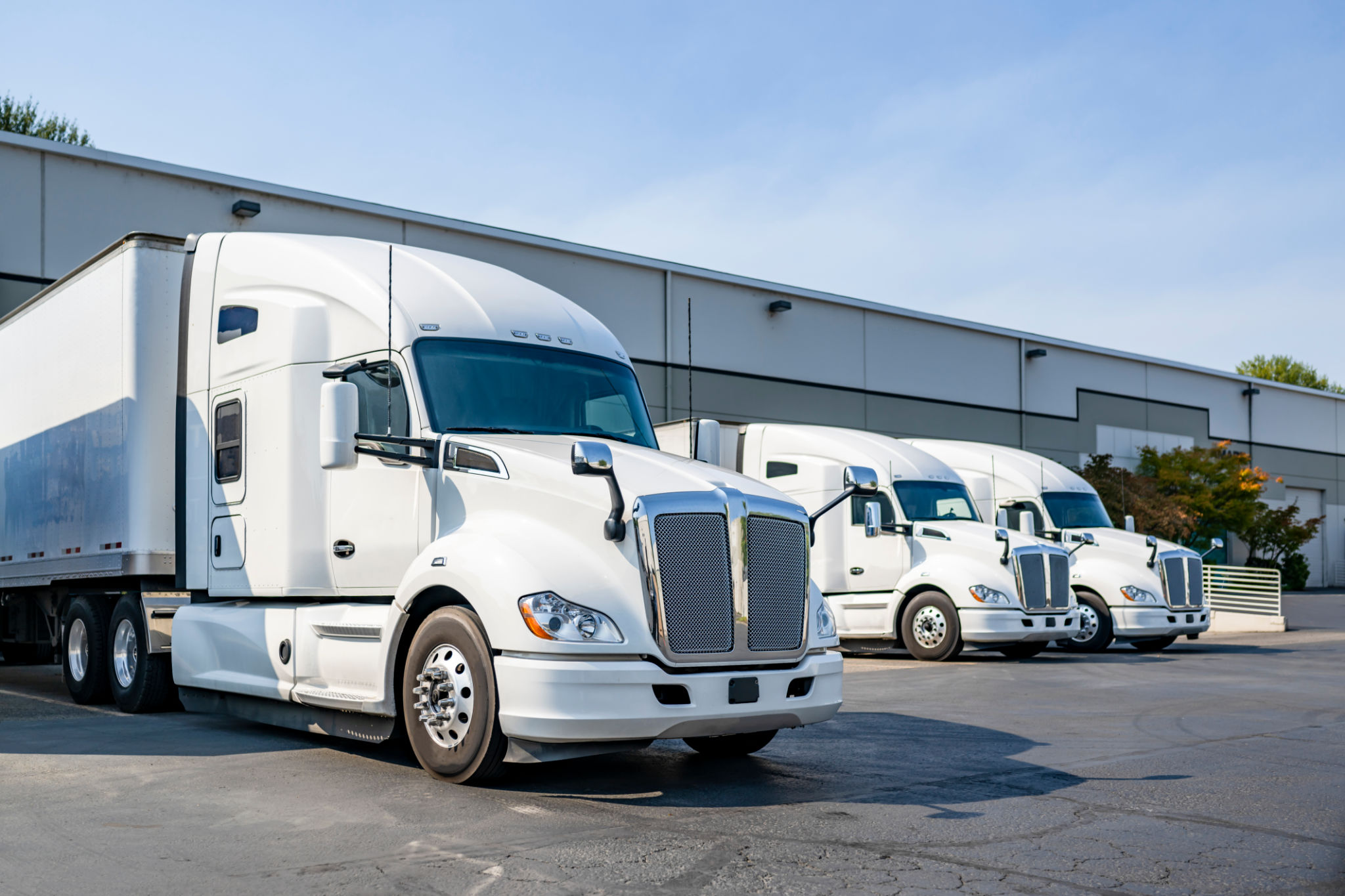 Row of the big rig semi trucks tractors with dry van semi trailers standing in warehouse docks loading cargo for the next delivery freight Row of the big rig semi trucks tractors with dry van semi trailers standing in warehouse docks loading cargo for the next delivery freight