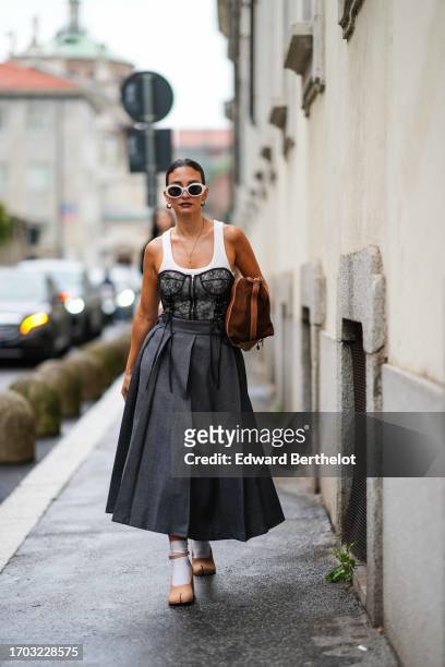 Guest wears earrings, white sunglasses, a white sleeveless top under a black lace corset, a grey pleated long skirt, split-toe ankle strap shoes, a...