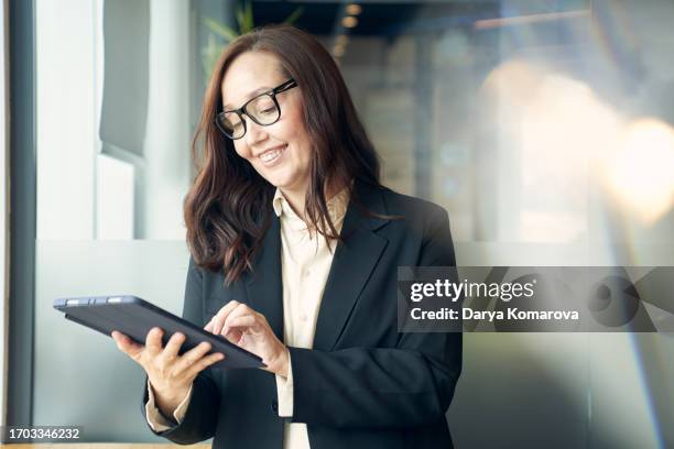 a woman with a digital tablet in a modern office. working from the office, a business woman in eyeglasses is touching the screen of the tablet and smiling. copy space and lens flare. - ceo stock pictures, royalty-free photos & images