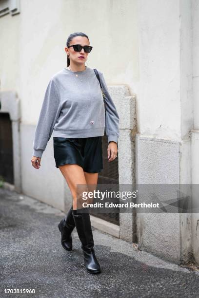 Guest wears earrings, sunglasses, a grey sweatshirt, a black leather mini skirt, black leather under-the-knee boots, outside Aigner, during the Milan...